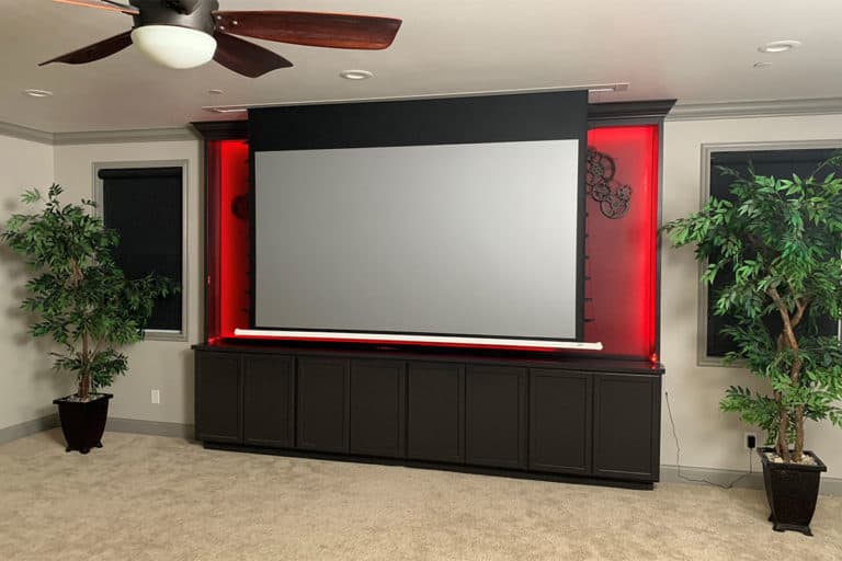 Large projection screen in a home theater with red accent lighting and surrounding potted plants.